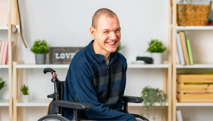 Smiling man in a wheelchair wearing a dark striped sweater, seated in a bright room with shelves and plants.