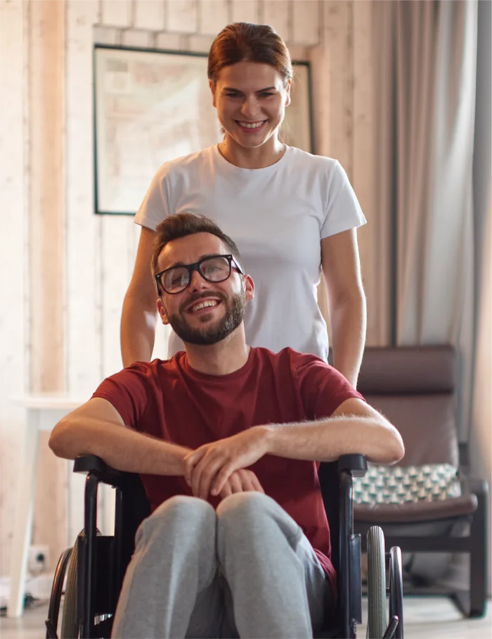 Smiling man in a wheelchair with a woman standing behind him in a cozy, softly lit indoor setting.